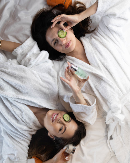 Two women in white robes lying on a bed with cucumbers on their eyes and skincare products.